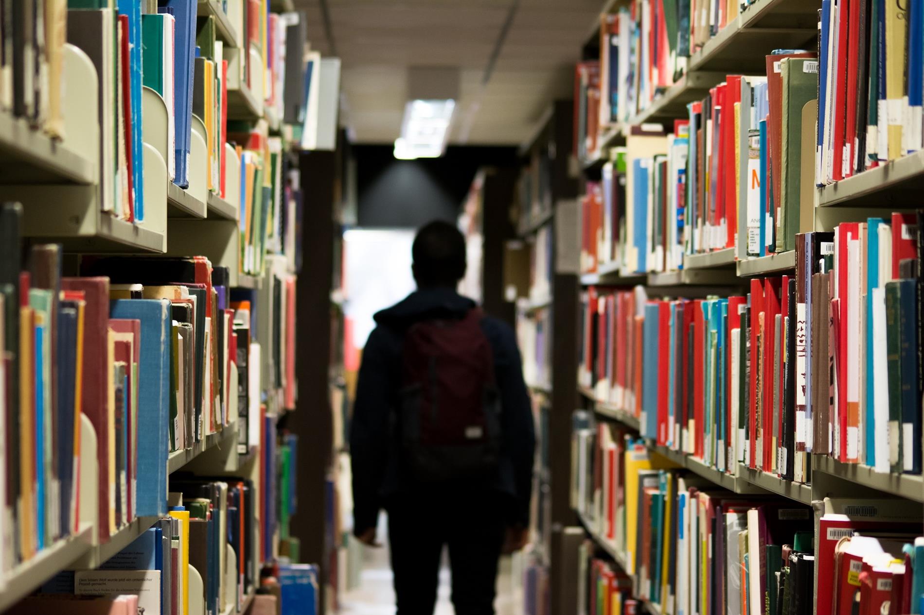 Silhouette of a person with a backpack walking away down the aisle between rows of books on library shelves