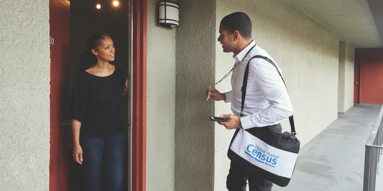 Census 2020 employee standing at door speaking to a person at their home. Census 2020 employee standing at door speaking to a person at their home.